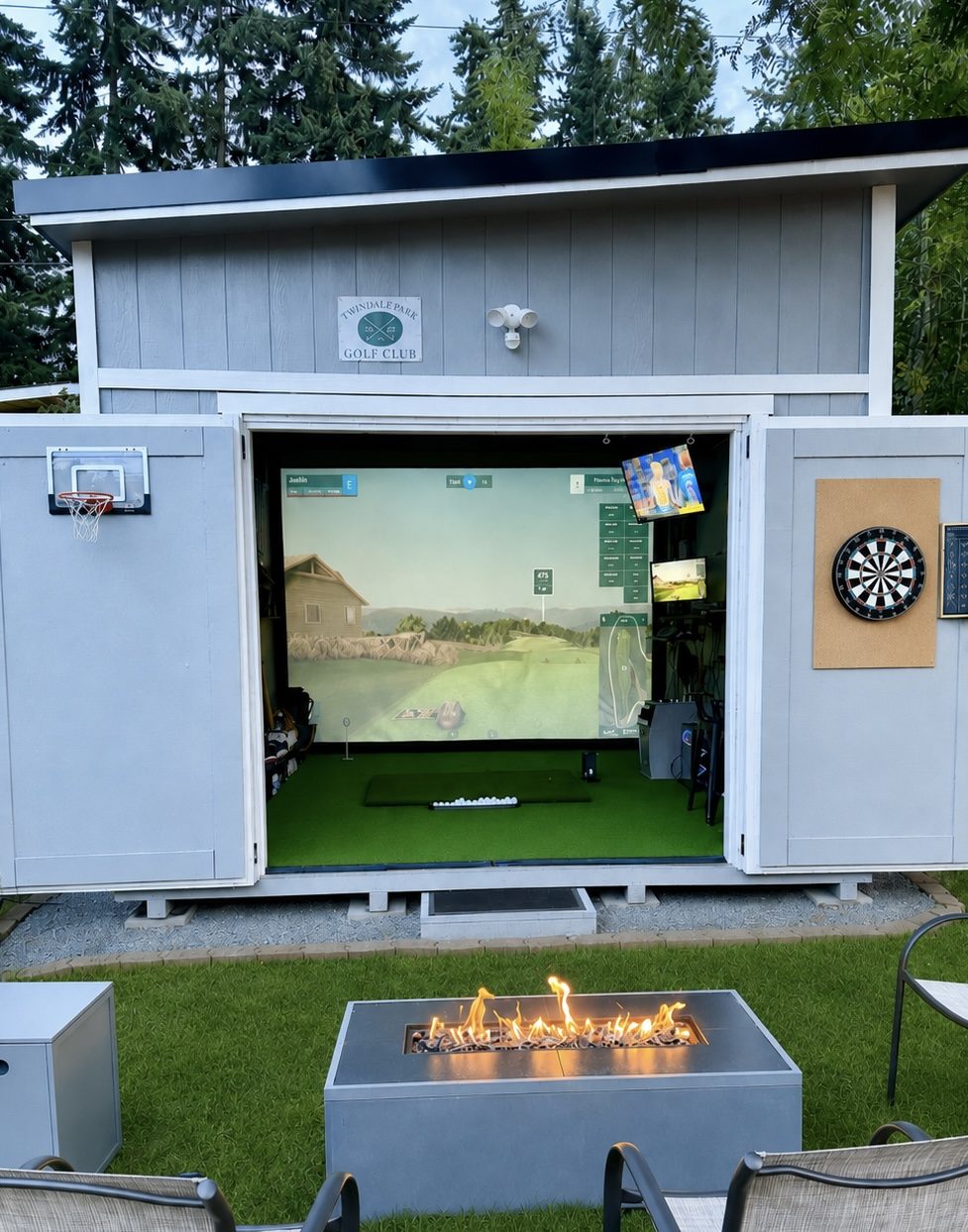A backyard golf simulator shed with its doors open, showing the simulator screen, hitting mat, a basketball hoop and dartboard on the flanking doors, and a fire pit out front.